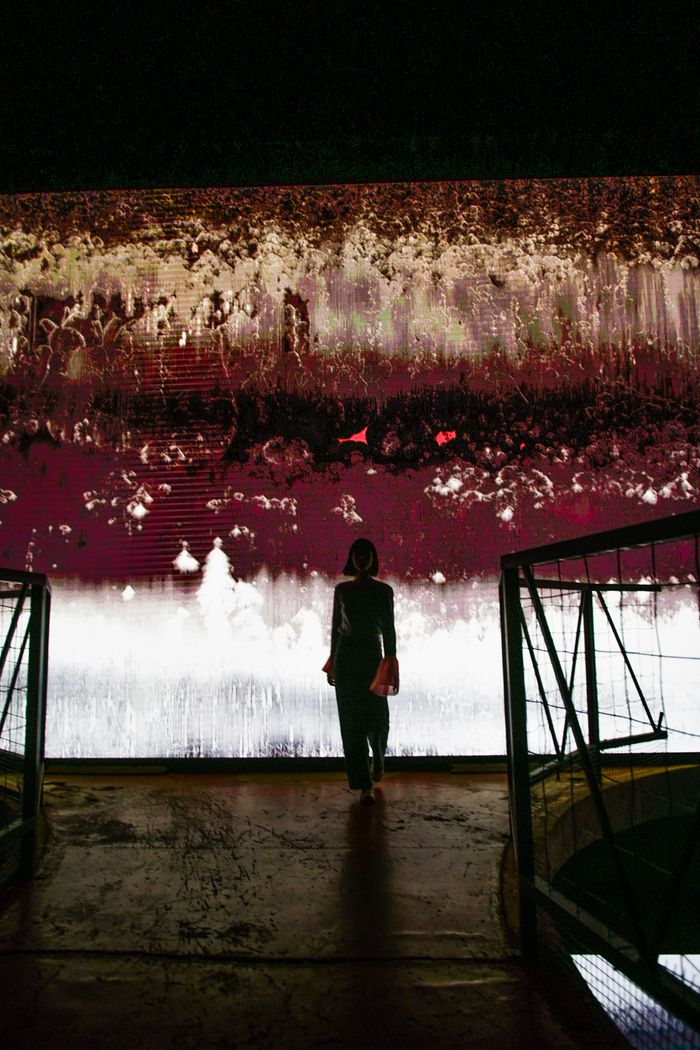 A person stands in front of a colorful abstract wall display in a dark indoor setting.